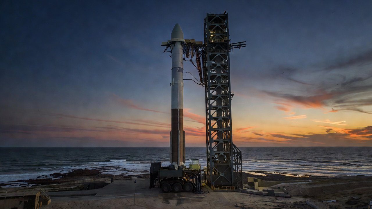 Large two-stage rocket standing on a coastal launch pad at dusk with service tower and mobile launch platform